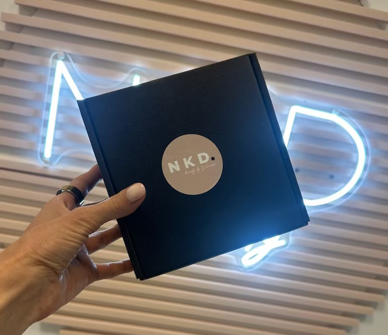 Hand holding a black box with 'NKD' logo against a decorative wall with neon lights