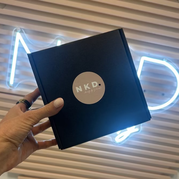 Hand holding a black box with 'NKD' logo against a decorative wall with neon lights